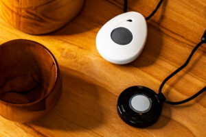 Black help button and white fall detection necklaces on a wooden table.