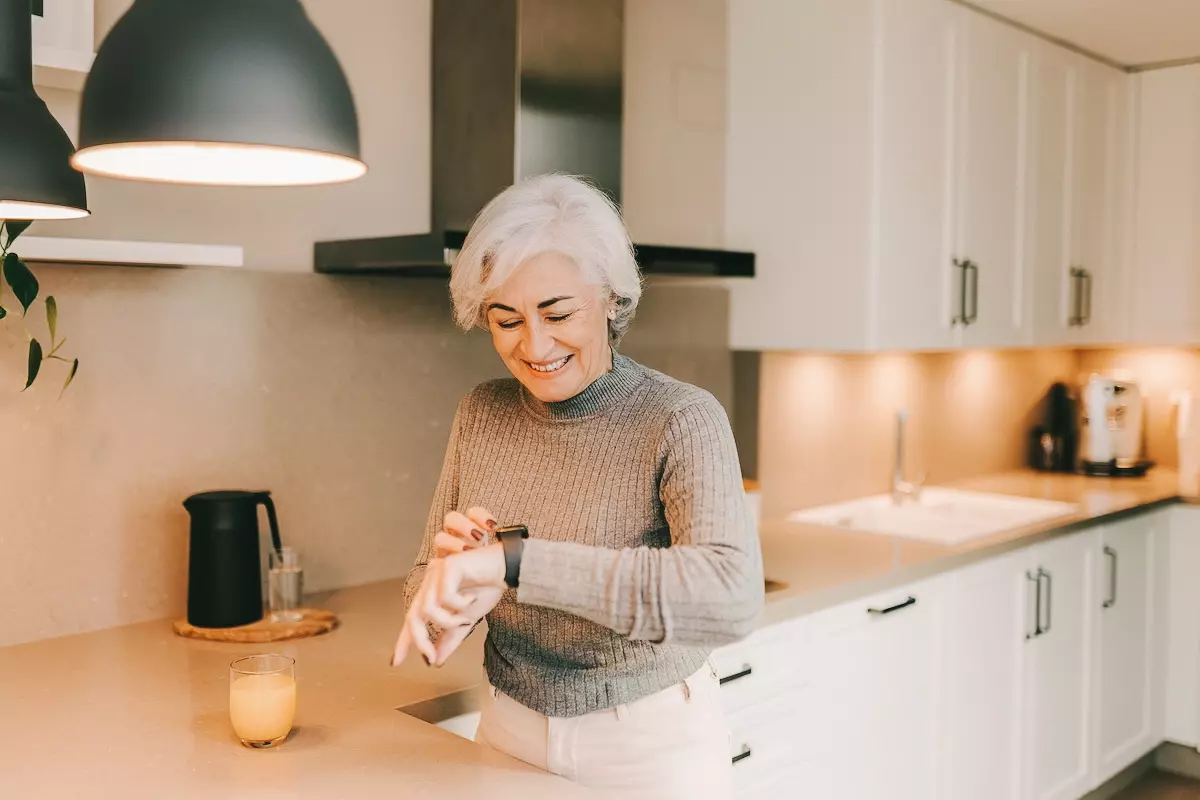Healthy senior woman smiling at her smartwatch while standing in her kitchen. Happy elderly woman monitoring her heart rate and fitness on a mobile app.