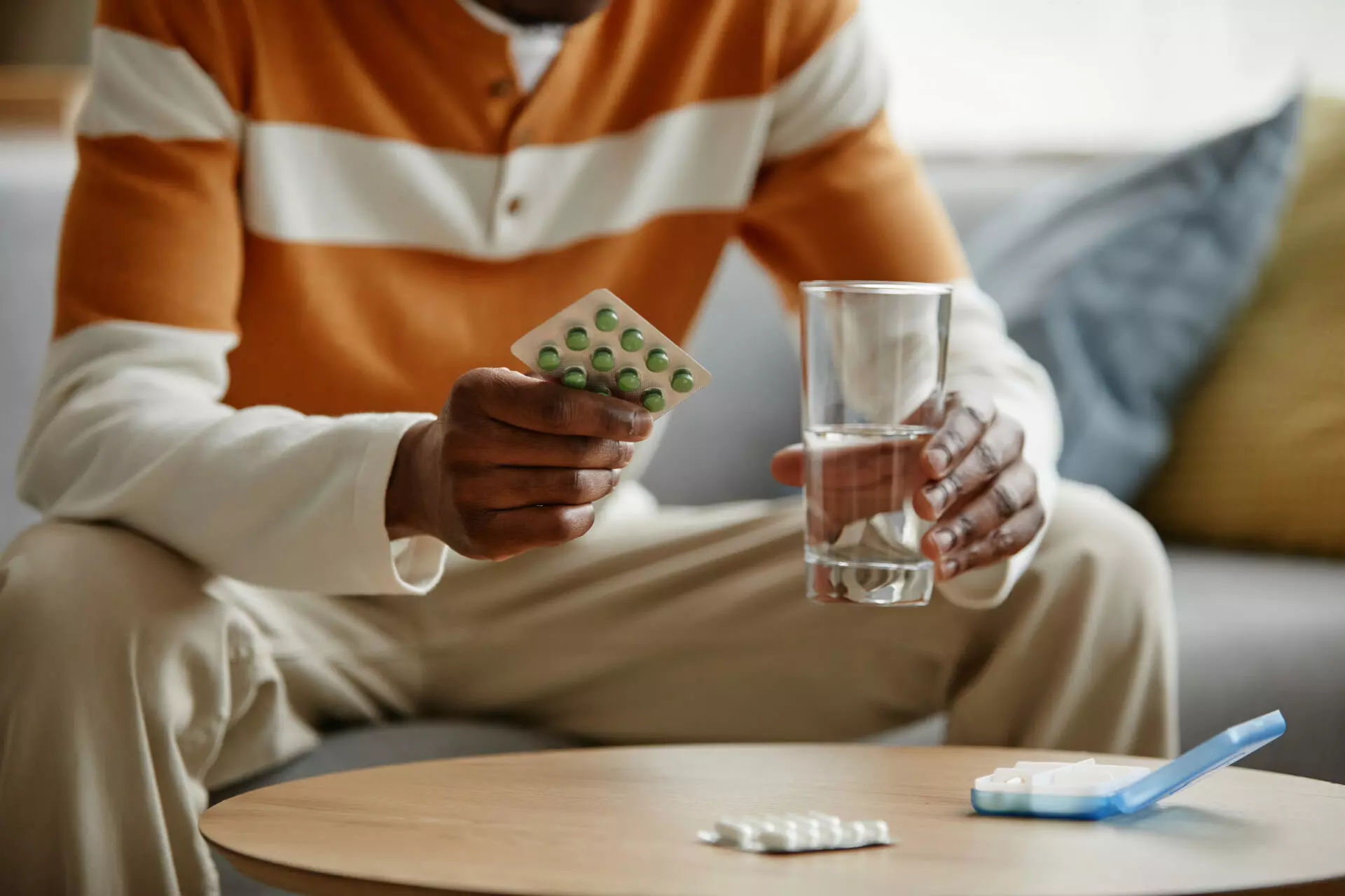 Close up of Black man holding blister of pills and glass of water while taking medication at home, copy space