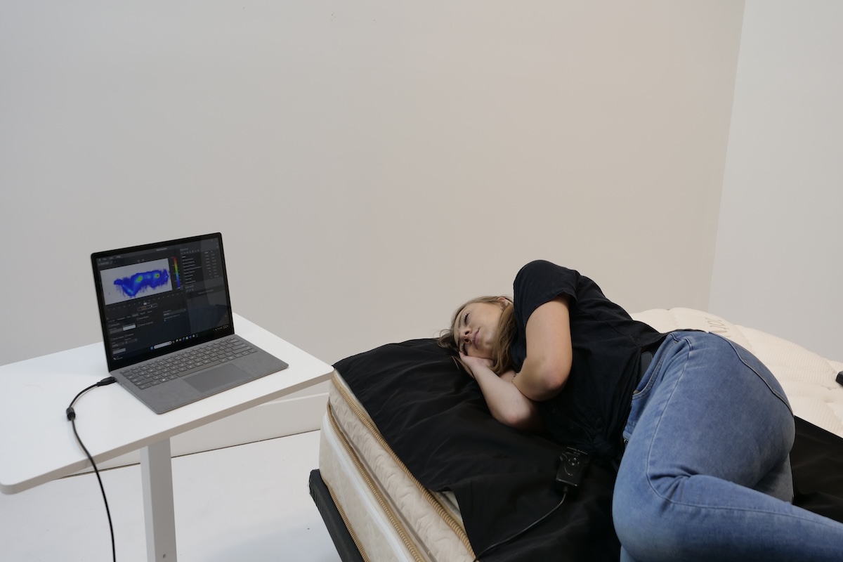 Woman lying on her side on top of a black mat on a mattress next to a laptop.