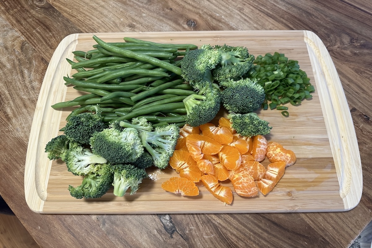 Green beans, broccoli, green onions, and mandarin oranges on a wooden cutting board.
