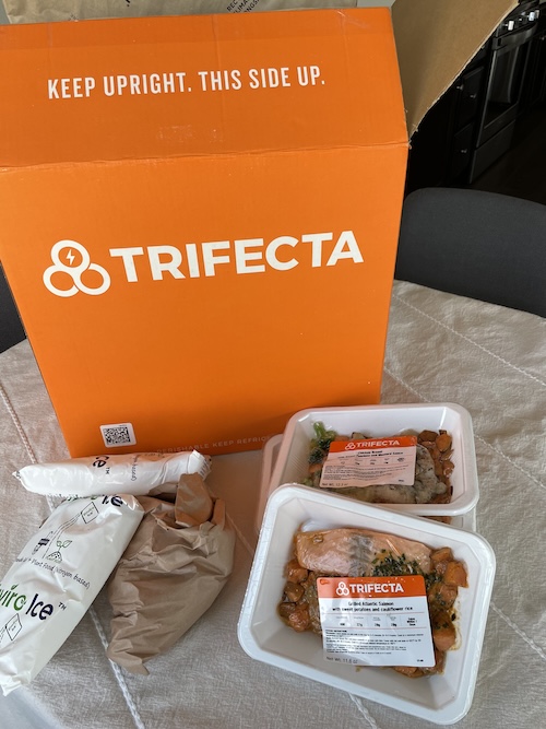 A Trifecta delivery box, ice packs, and packaged meals on a table. 