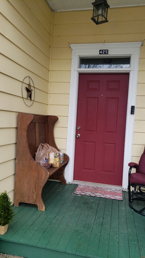 Green porch with red door and bagged grocery delivery on a wooden bench.