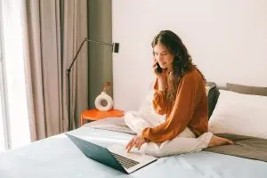 Woman sitting on her bed talking on the phone and looking at her laptop.