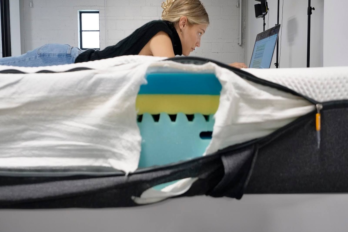 One of our testers lies on her stomach looking at a laptop screen on top of the Emma Original, with the side view showing an opening in the mattress and its interior composition. 