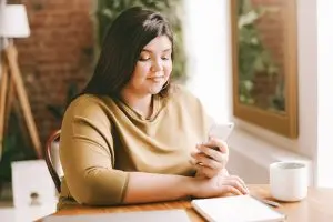 A young woman sits at a table in a brightly lit room while attending therapy sessions through her phone.