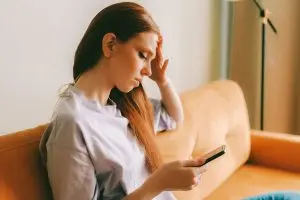 A young woman sitting on a yellow couch looking at mobile phone screen, holding her head.