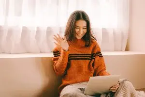 Young woman in an orange sweater sitting on a couch smiling and waving at a laptop screen.