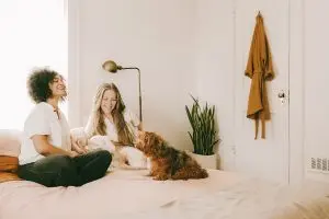 Two women and a small dog sit on top of a bed smiling.