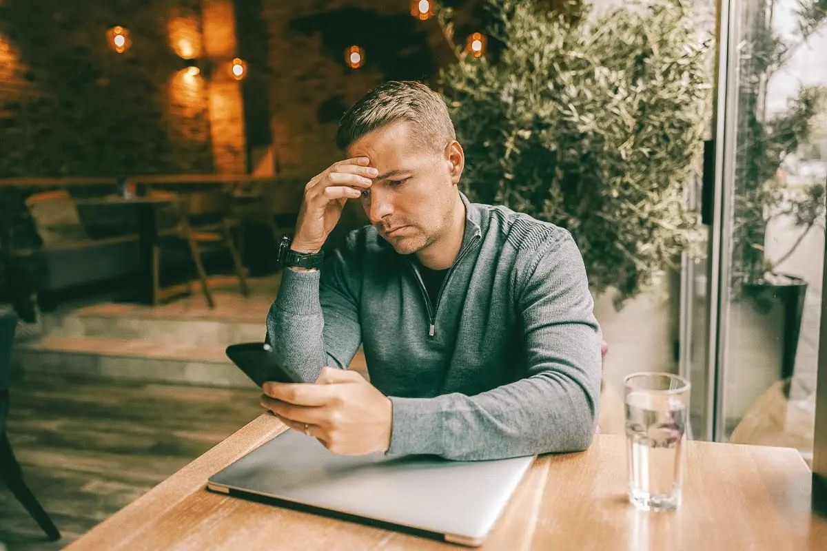 Worried man sitting at table with smartphone and laptop.