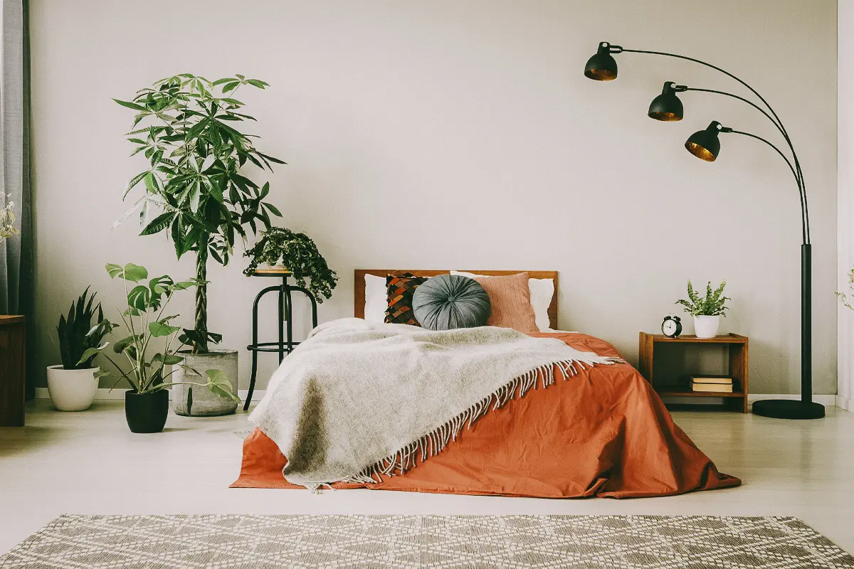 A bed with an orange comforter, a beige blanket, and three decorative pillows on top in a large bedroom with multiple potted plants and trees.