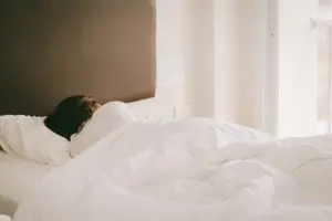 A woman sleeps with her back to the camera in a bed with a white comforter and sheets.